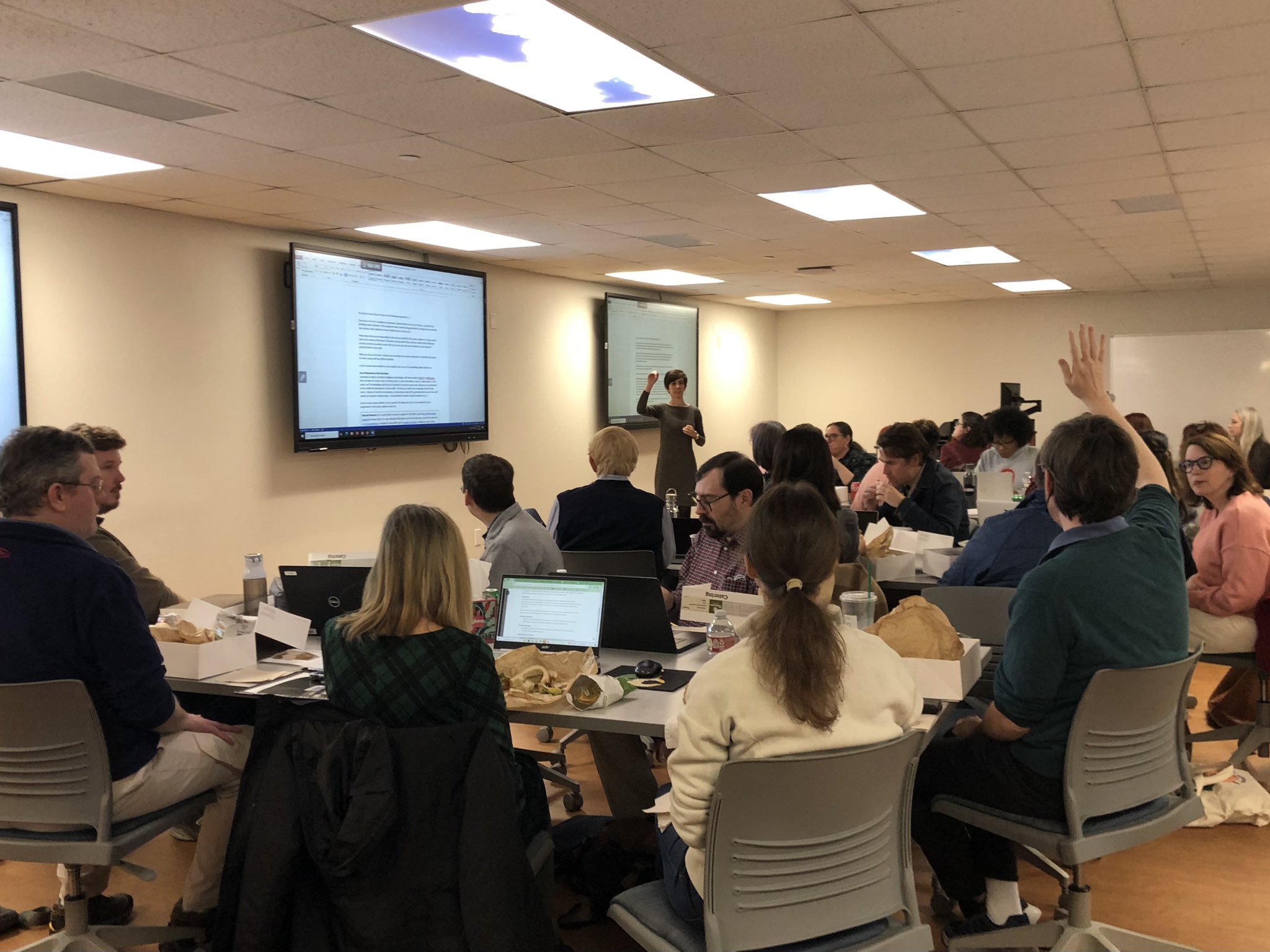 Photograph from the back of a full room of workshop participants. A woman stands at the front of the room by a projector screen, while a participant at the back raises his hand. 