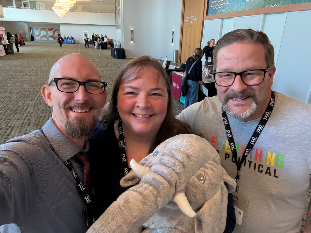 Three people pose for a selfie: two white men with glasses and a white woman holding a large elephant. In the background is a generic conference lobby area.
