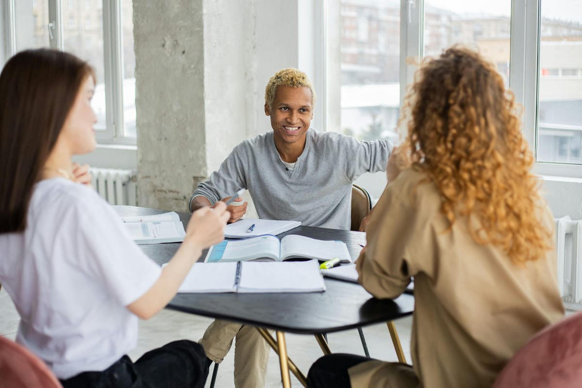 Three adults sit at a table in a white industrial space. They have books and notebooks open in front of them. No digital devices are in view.